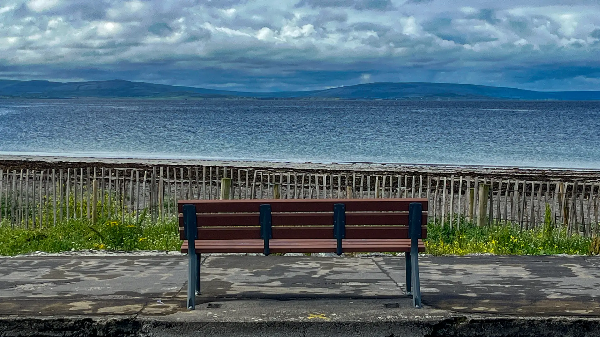 A seafront image with an empty bench facing towards the Burren in Co. Clare.
